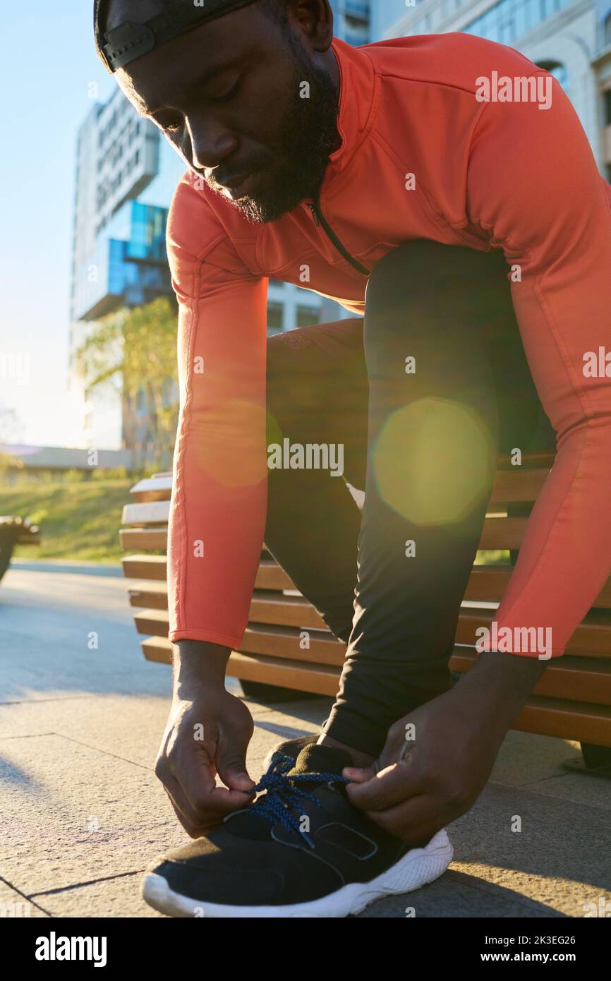 Giovane atleta maschile che lega la scarpa di sneaker mentre si siede sulla panca in ambiente urbano agter jogging e piegarsi sopra la gamba Foto Stock Giovane atleta maschile che lega la scarpa di sneaker mentre si siede sulla panca in ambiente urbano agter jogging e piegarsi sopra la gamba Foto Stock
