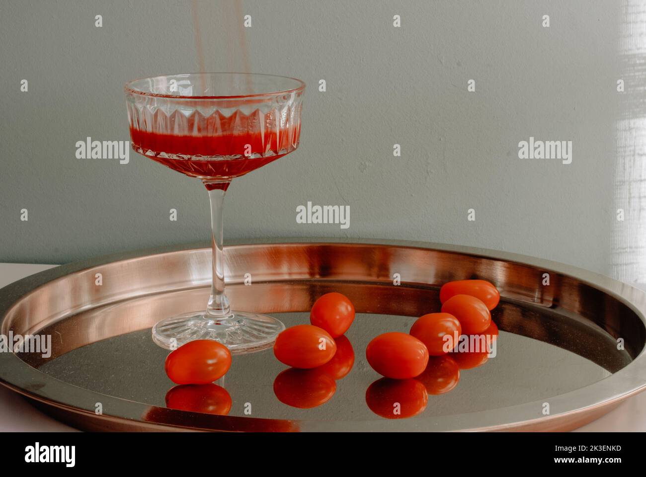 bel vetro di cristallo con pomodori rossi versando un fusto di pomodoro rosso si trova su un vassoio di metallo placcato oro illuminato dalla luce del sole che getta un'ombra su un Foto Stock