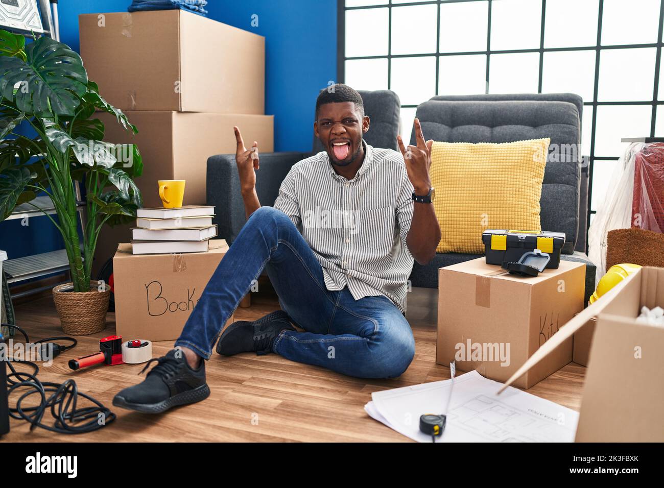 Uomo afroamericano seduto sul pavimento in una nuova casa gridando con espressione pazza facendo il simbolo del rock con le mani in su. Music star. Concetto di musica pesante. Foto Stock