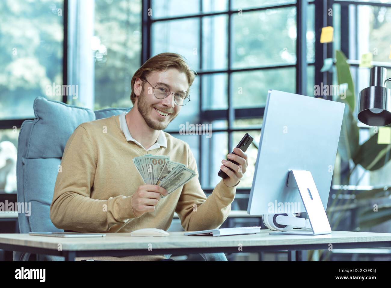 Ritratto di investitore finanziario di successo, uomo d'affari sorridente e guardando la macchina fotografica uomo in possesso di dollari in contanti e telefono cellulare che lavora all'interno di un ufficio moderno. Foto Stock