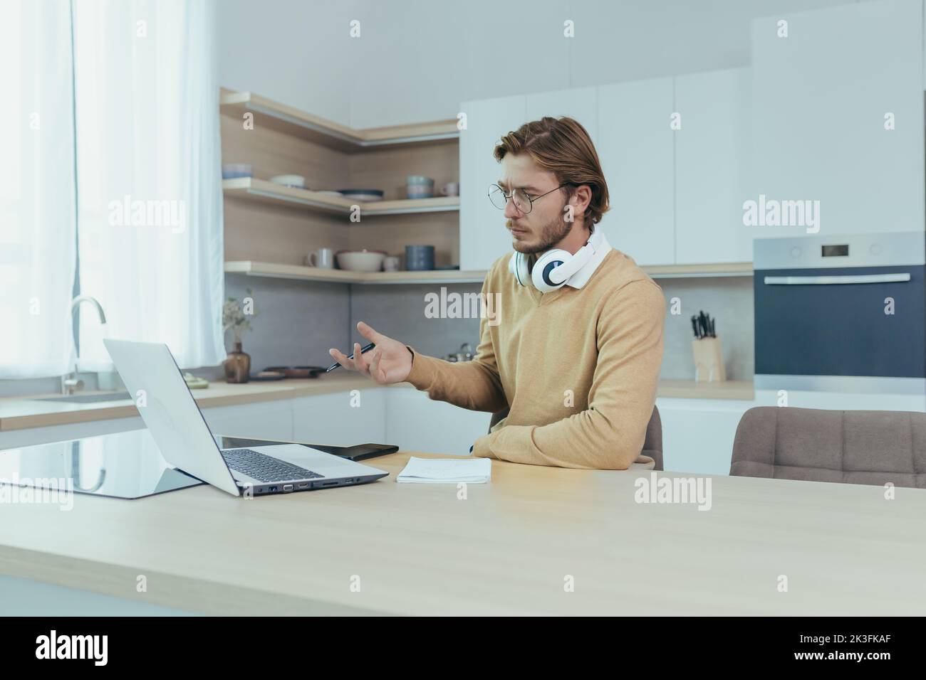 Giovane uomo d'affari che lavora a distanza da casa seduto in cucina, uomo che utilizza il computer portatile per videochiamare concentrandosi e parlando seriamente in una riunione online con i colleghi colleghi. Foto Stock