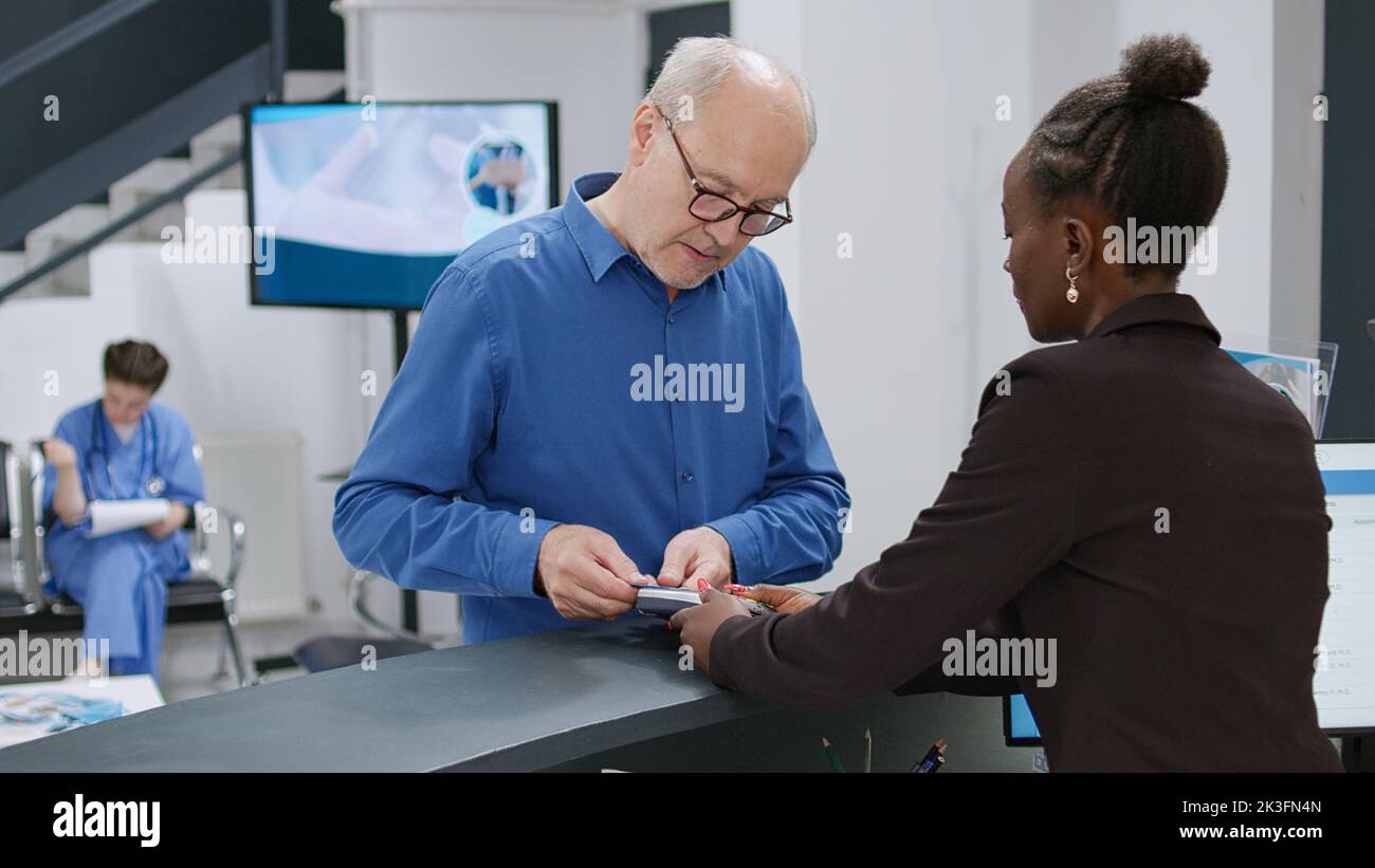 Senior man che paga con carta di credito alla reception dell'ospedale, effettuando una transazione di pagamento elettronico per appuntamento e trattamento di controllo medico. Addetto alla reception che aiuta l'uomo a pagare. Foto Stock