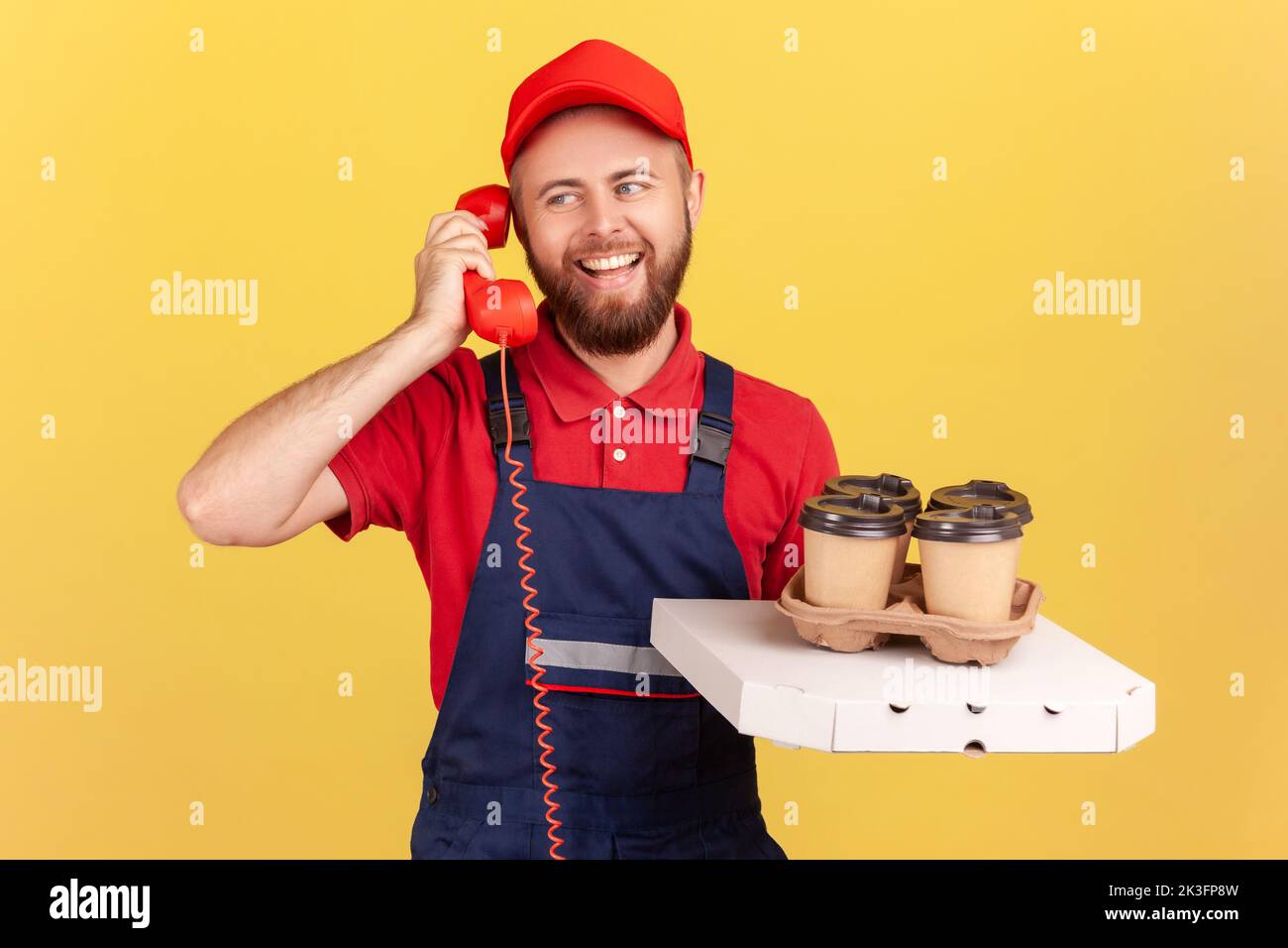 Ritratto di uomo corriere in tuta t-shirt rossa e cappello parlare telefono fisso e caffè con pizza, chiamata per ordinare fast food. Studio al coperto isolato su sfondo giallo. Foto Stock