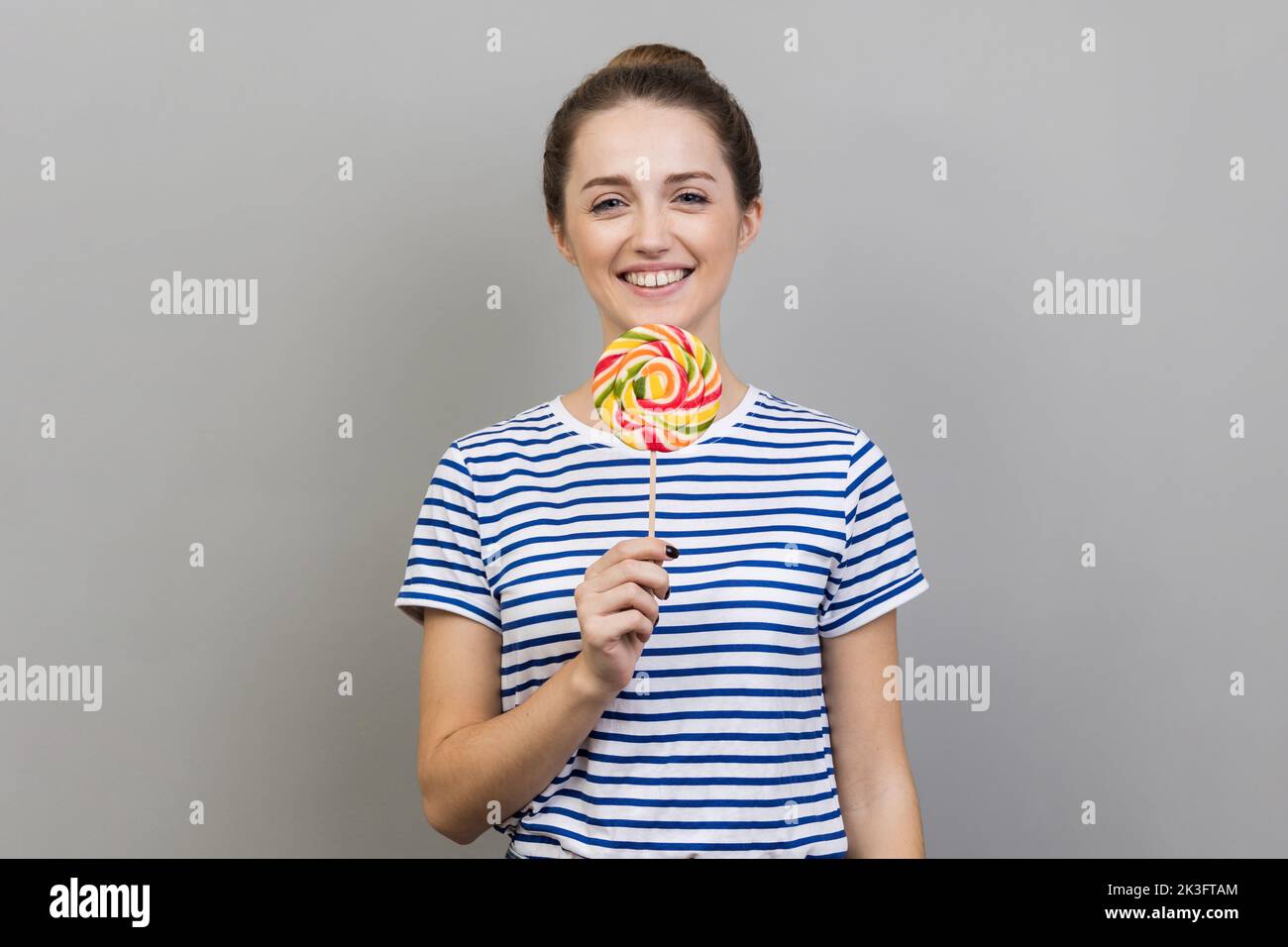 Ritratto di donna sorridente che indossa T-shirt a righe tenendo a righe colorato lollipop sul bastone in mano, pazzo di dolci dolciumi. Studio in interni isolato su sfondo grigio. Foto Stock