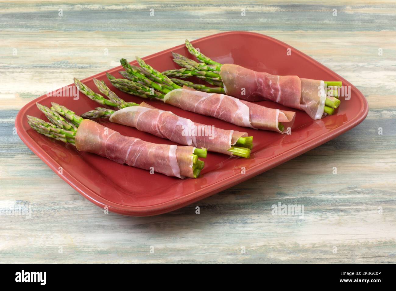 Grappoli di asparagi crudi avvolti in un piatto rosso a fette di prosciutto in preparazione alla cottura Foto Stock