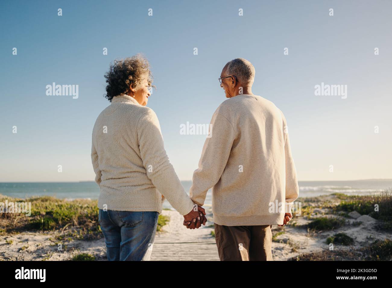 Vista posteriore di una coppia anziana felice che tiene le mani mentre cammina giù un ponticello del piede alla spiaggia. Romantica coppia anziana che prende una vacanza rinfrescante sul mare Foto Stock