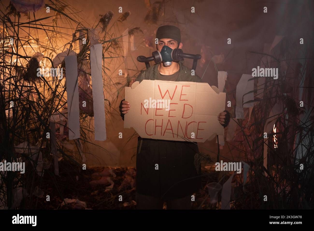 Un uomo in una maschera a gas con poster con le parole abbiamo bisogno di cambiare. Bruciare rifiuti di plastica sullo sfondo. Problemi ambientali: Rifiuti di plastica Foto Stock