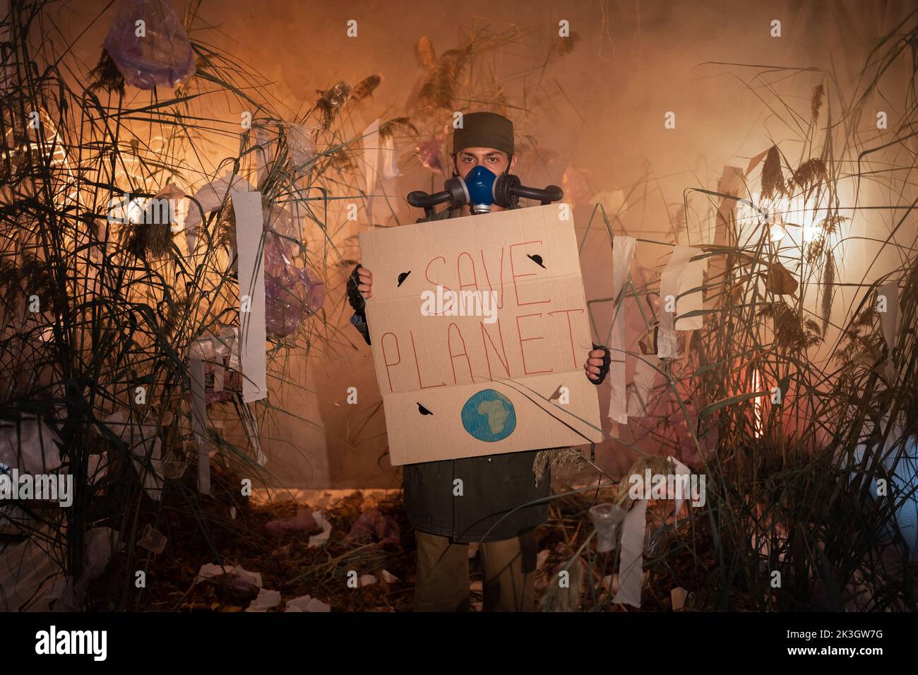 Un uomo in una maschera a gas con poster con le parole Salva pianeta. Bruciare rifiuti di plastica sullo sfondo. Problemi ambientali: Rifiuti di plastica, illegale Foto Stock