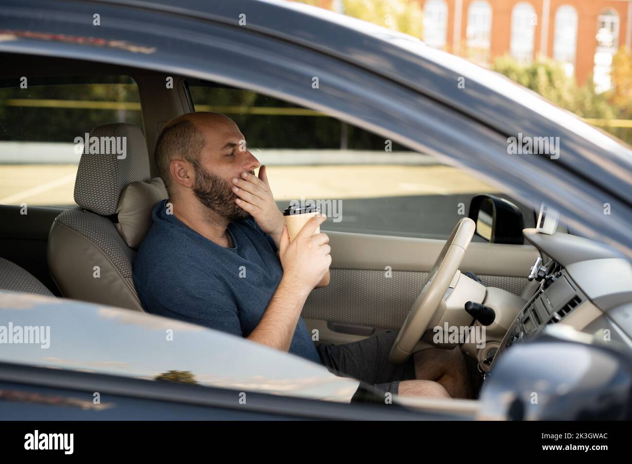 Uomo stanco che sbadigna sul sedile anteriore dell'auto che tiene il caffè per andare. Trasporto sonno privazione incidente Foto Stock