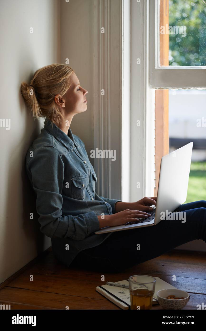 Pensando alla sua linea di apertura. Una giovane donna seduta su un pavimento che fa delle ricerche sul suo laptop e guarda fuori una finestra. Foto Stock