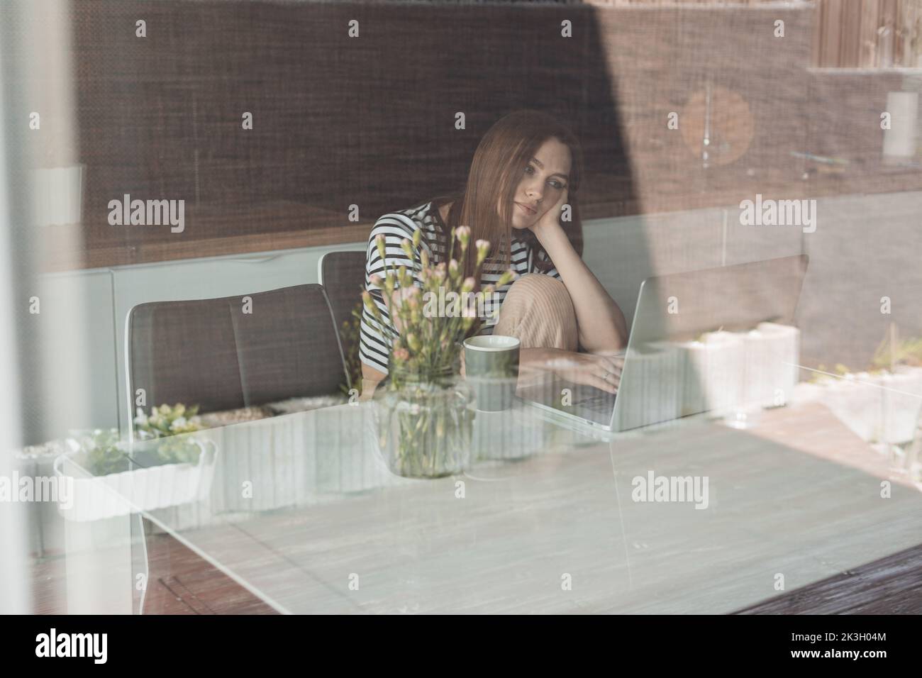 Donna depressa, triste, stanca dai capelli castani seduta in cucina, appoggiata a mano, che lavora su un computer portatile. Attraverso la vista finestra Foto Stock