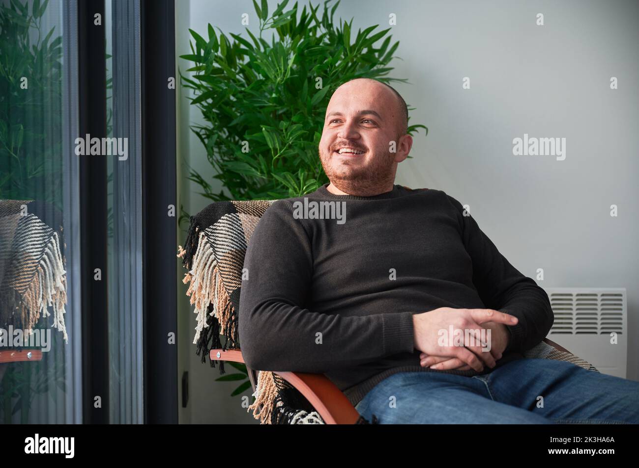 Uomo sorridente che si gode fine settimana all'interno di una casa fienile contemporanea. Buon turista maschile seduto su sedia in nuovo cottage. Foto Stock