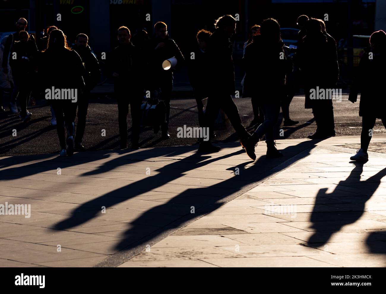 Una folla di persone in silhouette che attraversano la strada creando lunghe ombre.Back illuminato sole dà un'atmosfera drammatica per la fotografia Foto Stock