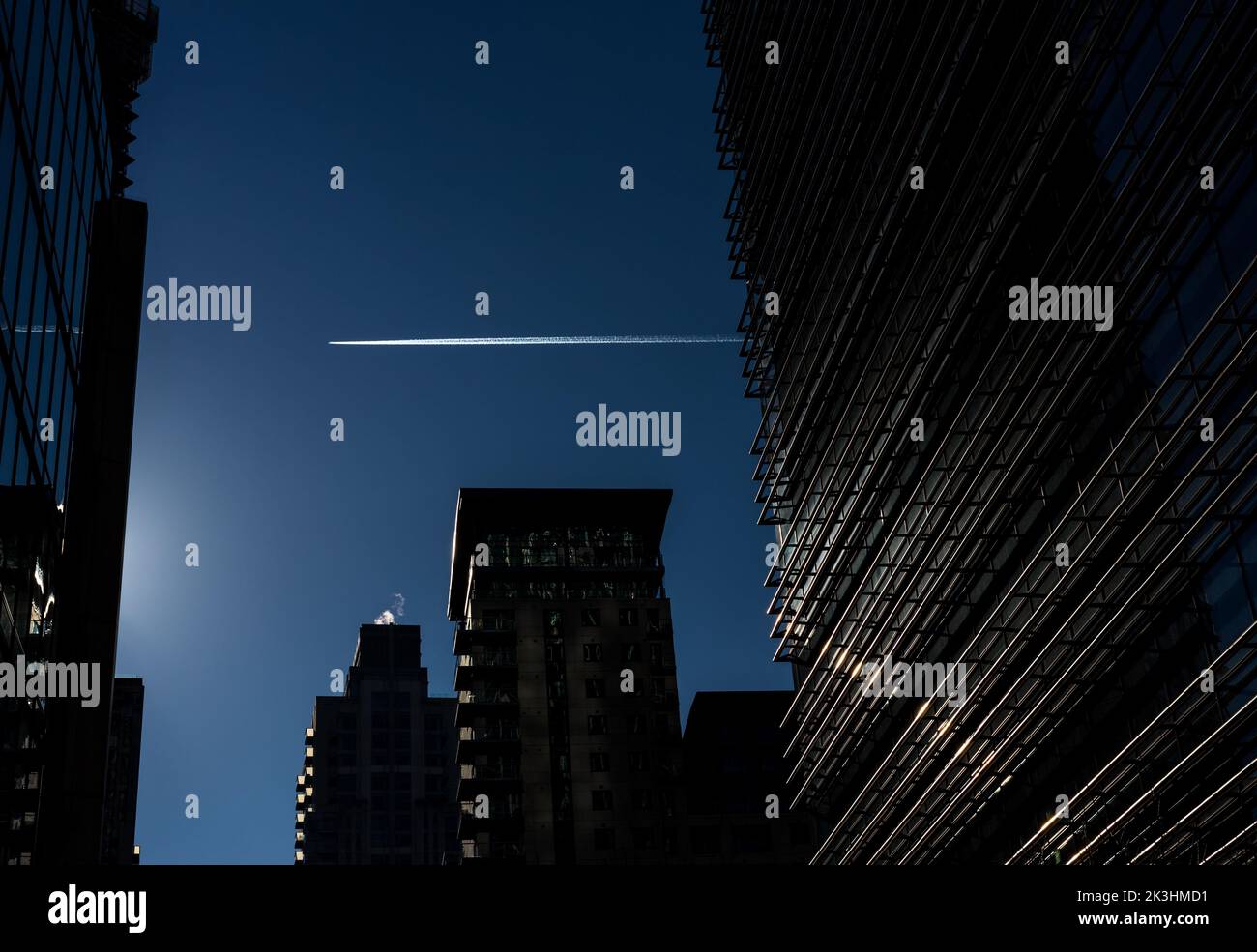 L'immagine del molo dei Canarie mostra alti edifici di vetro in silhouette contro un cielo blu scuro.Un aereo a getto distante lascia un percorso di scarico bianco. Foto Stock