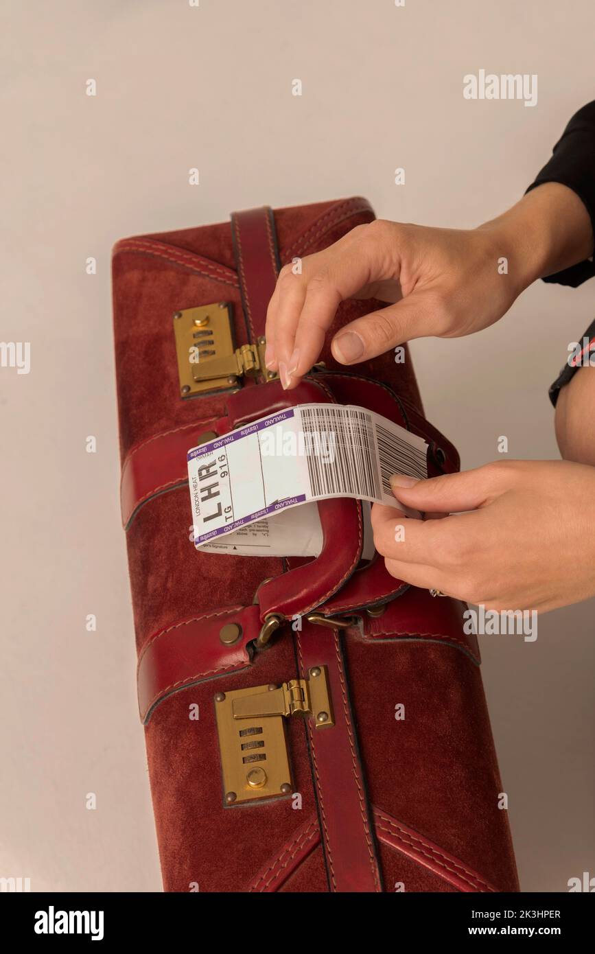 Enngland, Regno Unito. 2022. L'etichetta di identità della destinazione dei bagagli aerei viene attaccata a una maniglia della valigia in pelle rosso scuro dalle mani delle donne. Foto Stock