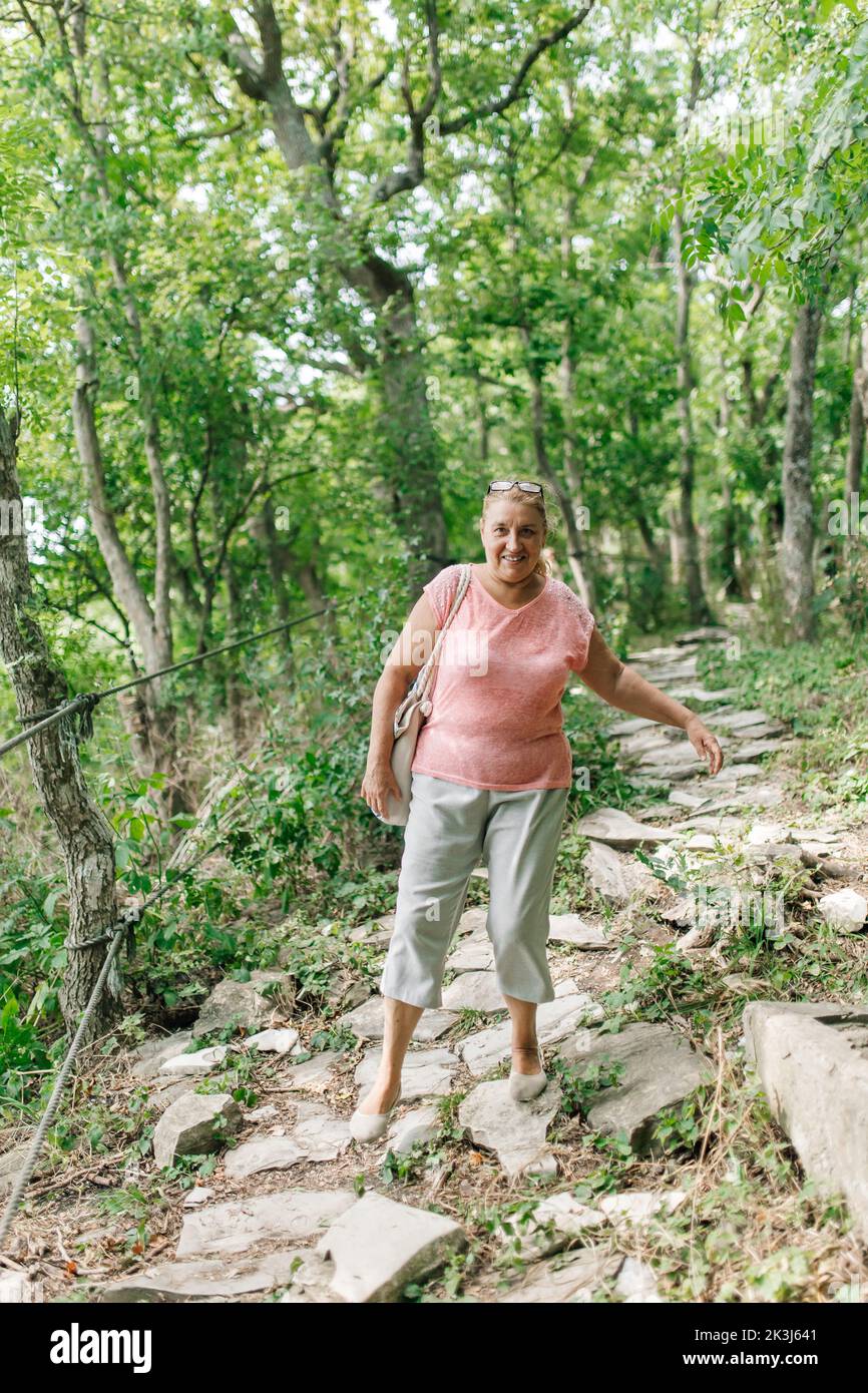 Donna di mezza età va singolarmente giù sentiero di pietra senza guardare i piedi nella foresta verde. Turistico con interesse a piedi lungo percorso pericoloso. Escursioni Foto Stock