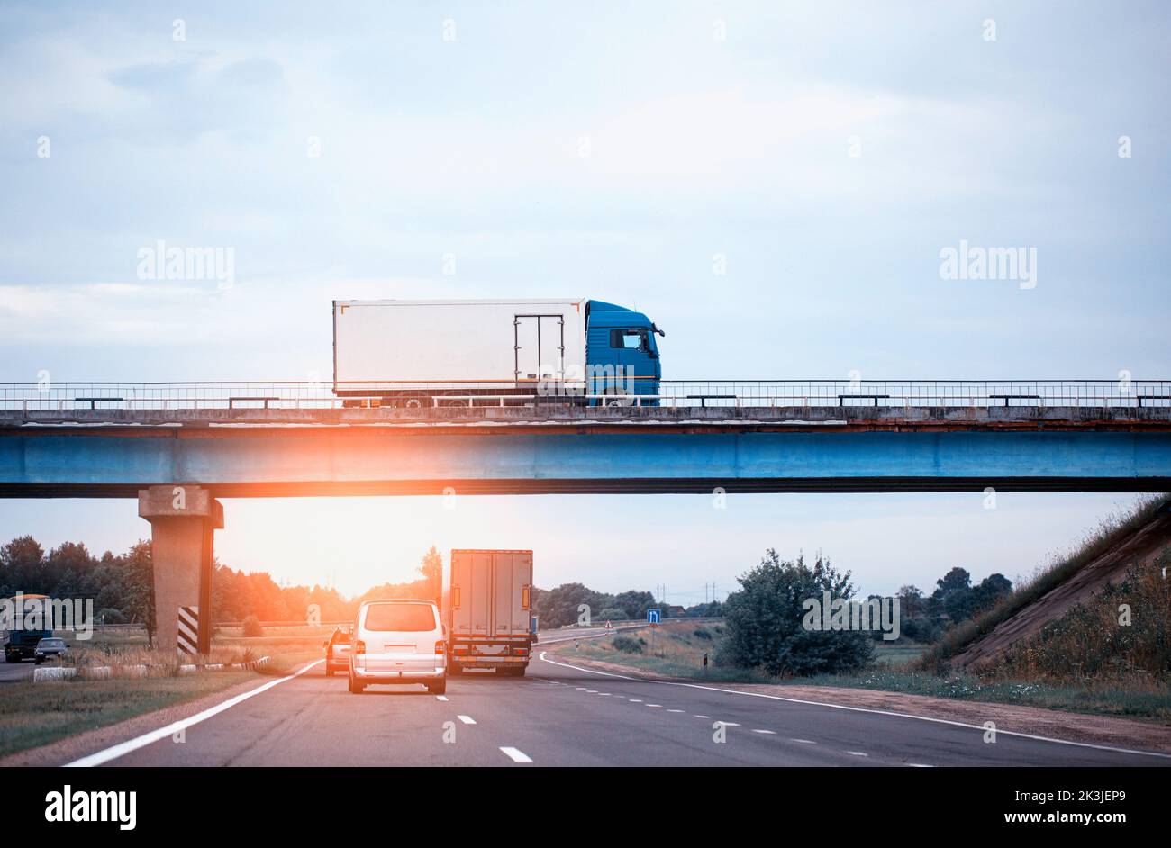 Traffico da camion e auto sulla strada. Un camion guida su un ponte sullo sfondo di un tramonto serale e cielo blu. Il concetto di importazione A. Foto Stock