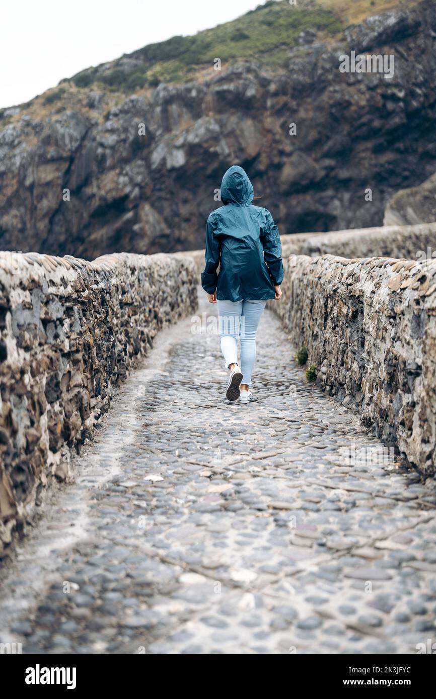 donna che indossa un impermeabile che cammina lungo una strada asfaltata racchiusa da due pareti di pietra Foto Stock