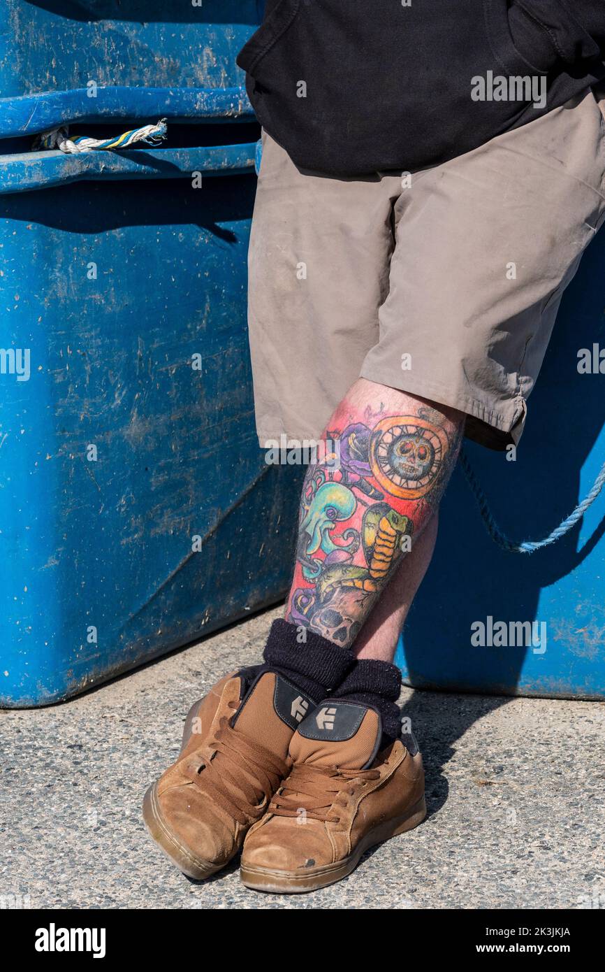 Un uomo che si rilassa contro i bidoni di stoccaggio con le gambe tatuate incrociate. Foto Stock