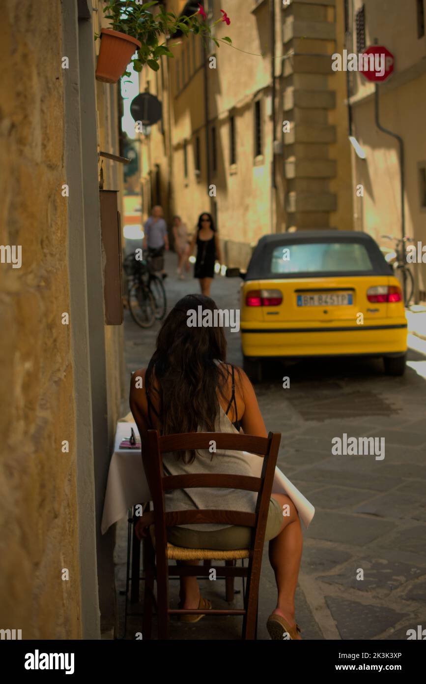 Donna dai capelli castani seduta da sola in un tavolo mentre un cabriolet giallo passa a Firenze Foto Stock