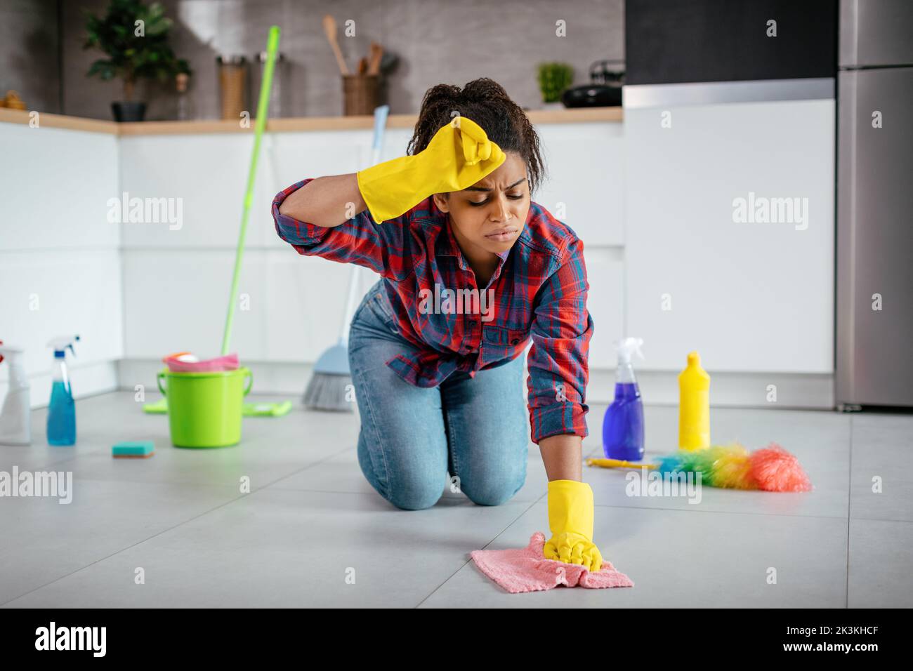 Stanco infelice giovane signora nera lava pavimento con i rifornimenti di pulizia, asciuga il sudore dalla fronte Foto Stock