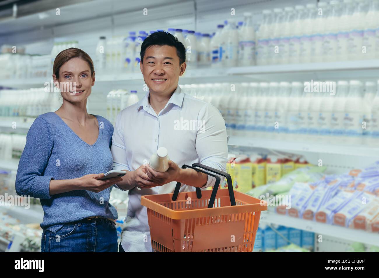 Shopping per tutta la famiglia. Ritratto di una giovane coppia interrazziale, di un uomo asiatico e di una donna in piedi in un supermercato nel reparto caseario, tenendo un cestino e una bottiglia di latte. Guarda la fotocamera. Foto Stock