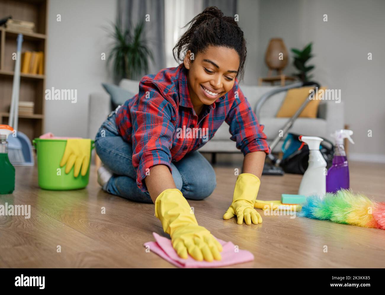 Felice giovane donna nera in guanti di gomma lava pavimento con i rifornimenti di pulizia, ama la pulizia in soggiorno Foto Stock