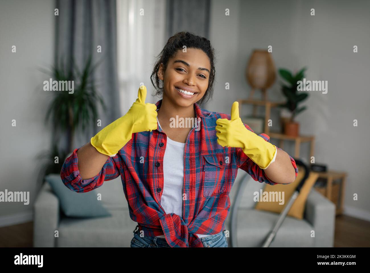 Ottimo lavoro. Felice giovane signora nera in guanti mostra pollici in su, gode di risultato di pulizia in salotto interno Foto Stock