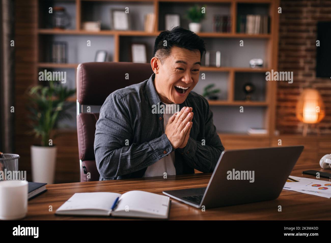 Uomo giapponese adulto felice guardare il portatile, gesturing e saluto, hanno videochiamata con il cliente Foto Stock