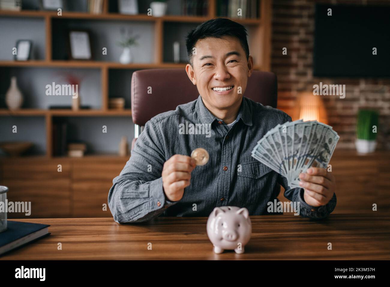 Soddisfatto successo maturo coreano mostra moneta bitcoin e molti dollari a tavola con la banca piggy Foto Stock