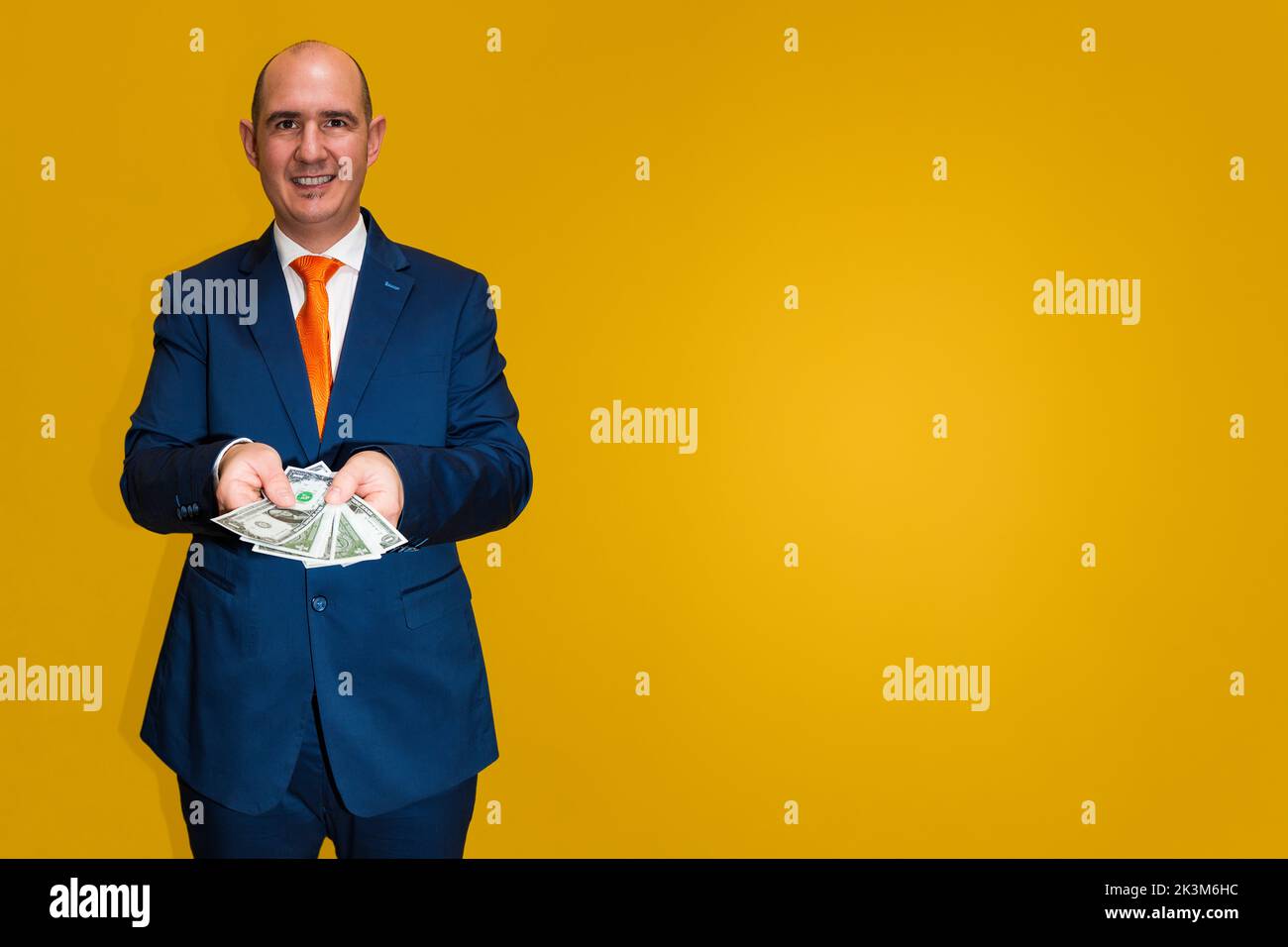 Un maschio calvo e caucasico adulta vestito con un vestito blu, una camicia bianca e una cravatta arancione sta tenendo diverse banconote da un dollaro con entrambe le mani. Lo sfondo è Foto Stock