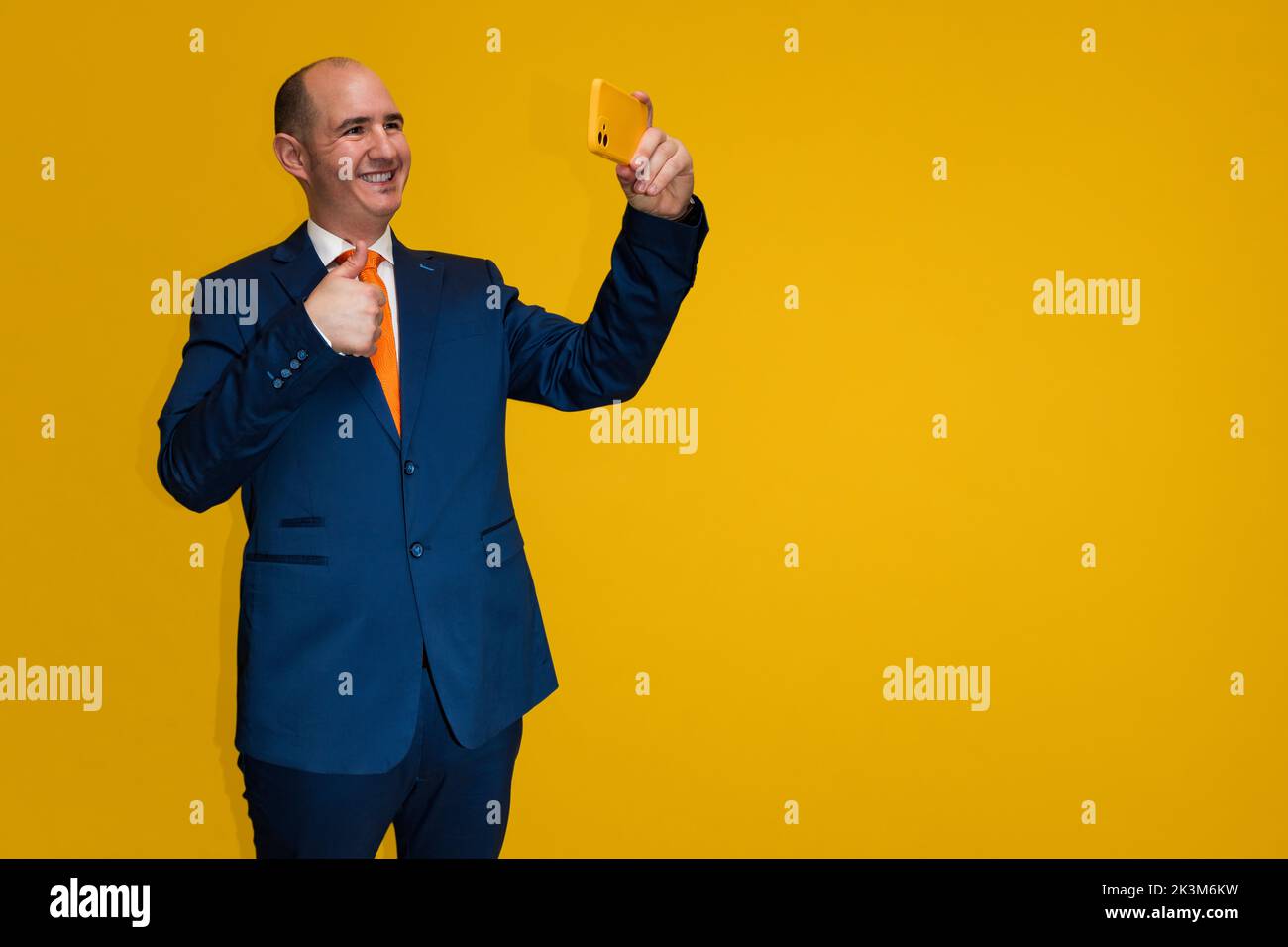 Un uomo adulto caucasico sorridente calvo che indossa un vestito blu, una camicia bianca e una cravatta arancione sta prendendo un selfie con una macchina fotografica del telefono delle cellule. Lo sfondo è yel Foto Stock