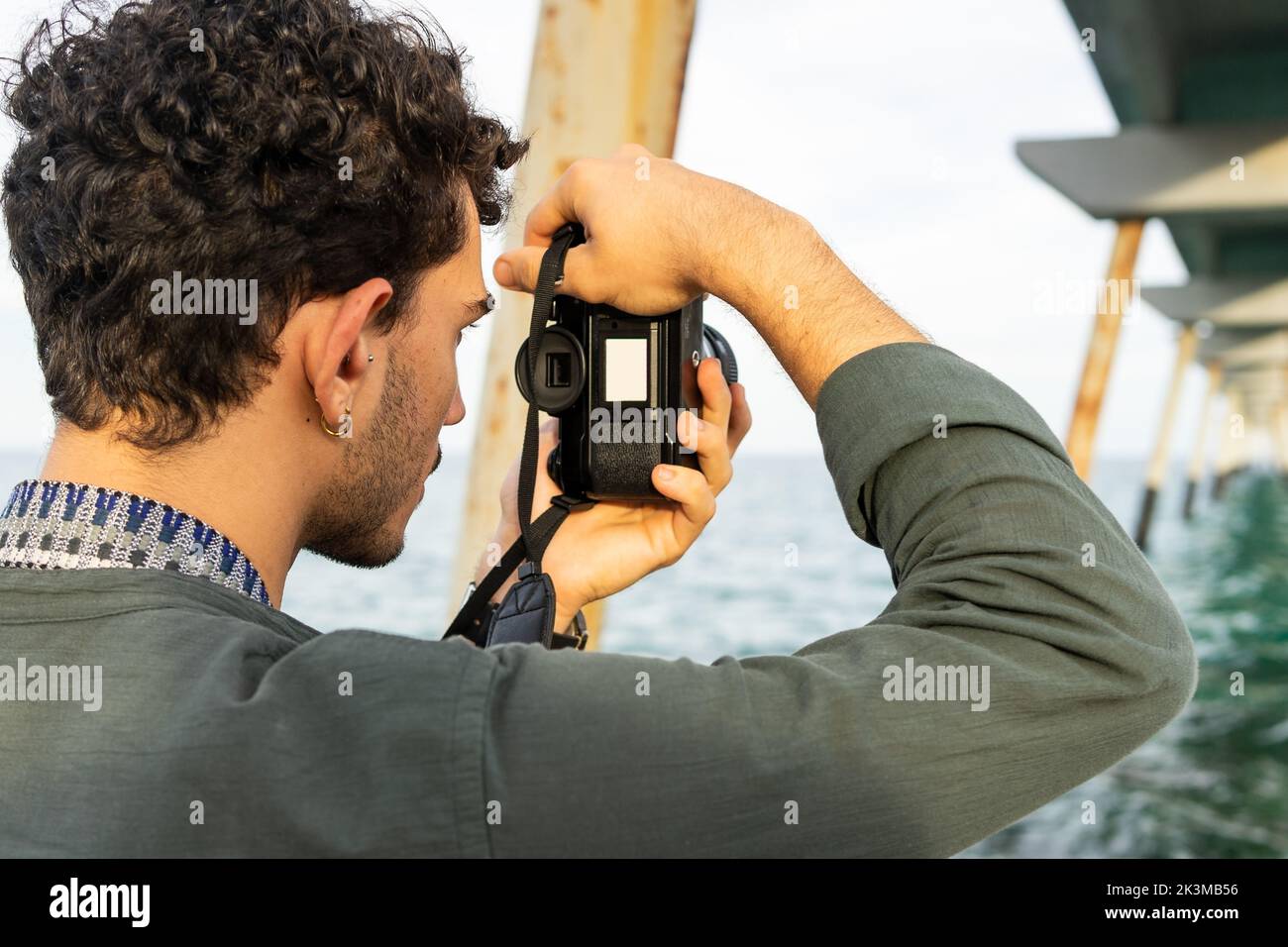 Irriconoscibile fotografo maschile che scatta ponti arrugginiti in acqua di mare mentre trascorri una nuvolosa giornata estiva in spiaggia Foto Stock