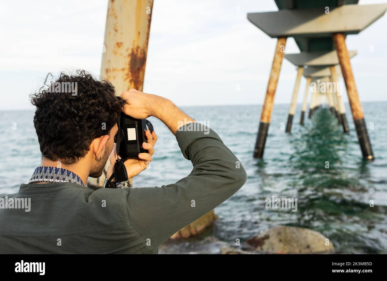 Irriconoscibile fotografo maschile che scatta ponti arrugginiti in acqua di mare mentre trascorri una nuvolosa giornata estiva in spiaggia Foto Stock