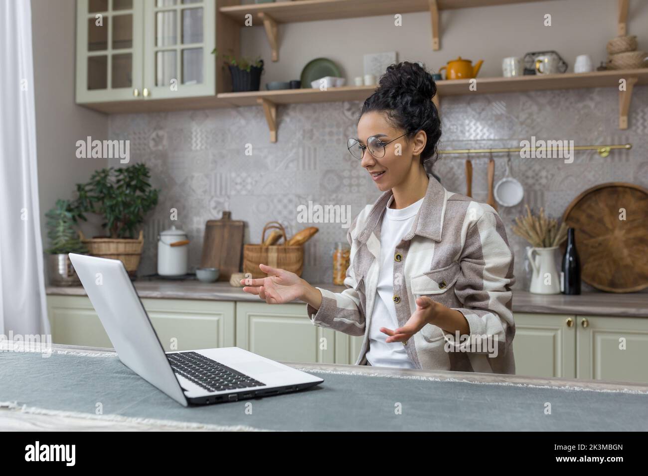 Giovane donna bella in occhiali e capelli ricci parlando in videochiamata, donna ispanica utilizzando il laptop per la comunicazione a distanza a casa in cucina. Foto Stock