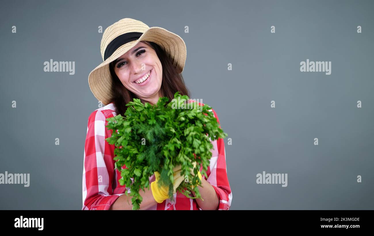 ritratto di donna sorridente contadina in camicia a pipa, guanti e cappello, odora un mazzo di prezzemolo fresco verde, verdi, su sfondo grigio, in studio. Cibo sano al vostro tavolo. Foto di alta qualità Foto Stock