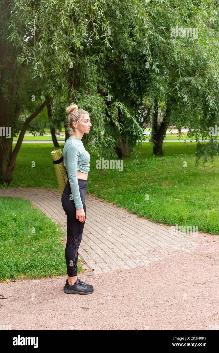 Una donna snella in abbigliamento sportivo, in piedi d'estate nel parco, con un tappetino verde da palestra per lo sport. Foto Stock