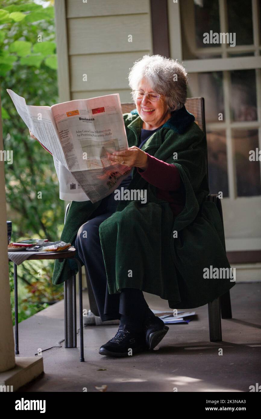 Una donna anziana felice dai capelli grigi siede sul portico leggendo il giornale Foto Stock