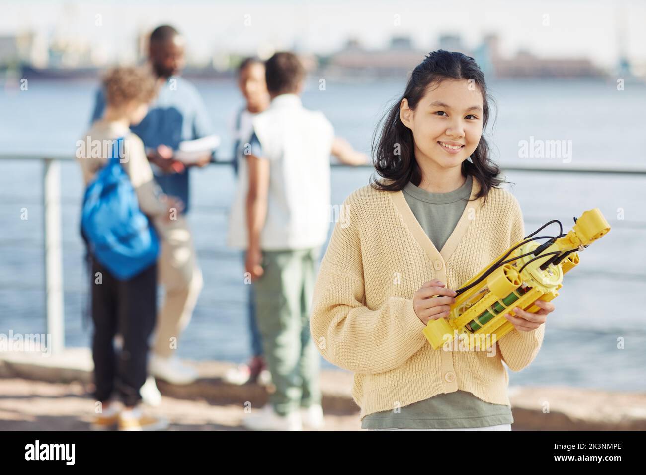 Girovita in su ritratto della ragazza asiatica adolescente che tiene modello di robot e sorridente alla macchina fotografica durante la lezione di ingegneria all'aperto con i bambini in background, copia spazio Foto Stock