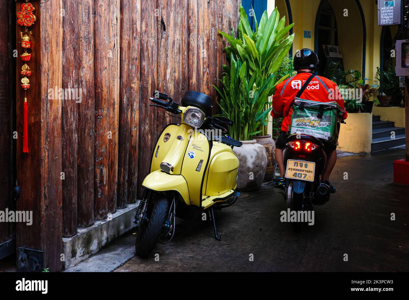 Bangkok, Thailandia - 23 gennaio 2022: Messaggero camicia arancione sta guidando la moto attraverso una piccola strada pedonale con il classico giallo scooter parcheggio ne Foto Stock