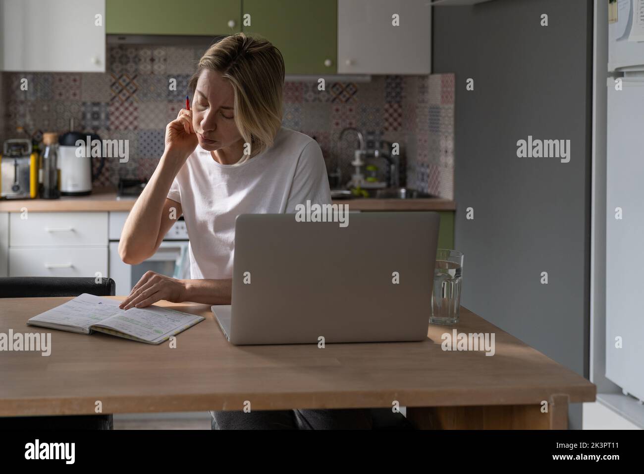 Una donna di mezza età affollata fa segni nel notebook e cerca un lavoro pagato tramite un computer portatile in cucina. Foto Stock