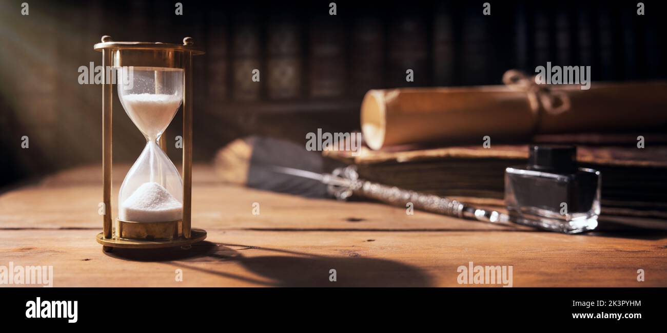 sabbia che scorre attraverso la clessidra su vecchio tavolo di legno con libri d'epoca e penna di penna. il tempo sta ticchettando. banner Foto Stock