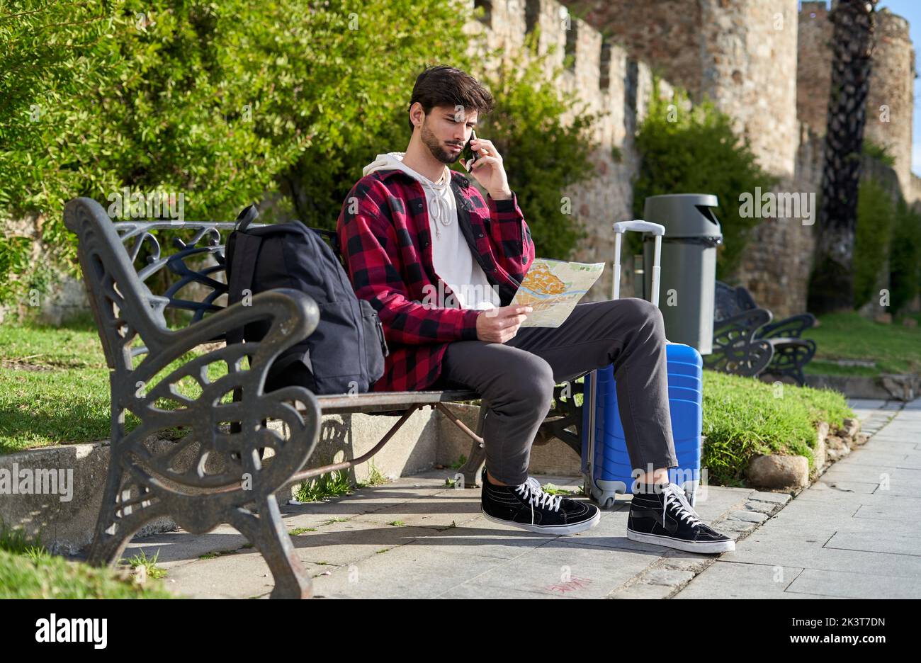 Corpo pieno di maschio barbuto turistico che controlla la mappa di carta e che parla sul cellulare mentre si siede sulla panca con lo zaino vicino all'edificio vecchio Foto Stock