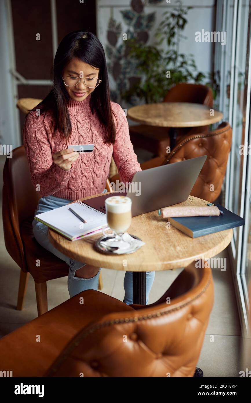 Una ragazza asiatica seduta nella caffetteria e utilizzando la sua carta di credito per lo shopping online sul computer portatile. Foto Stock