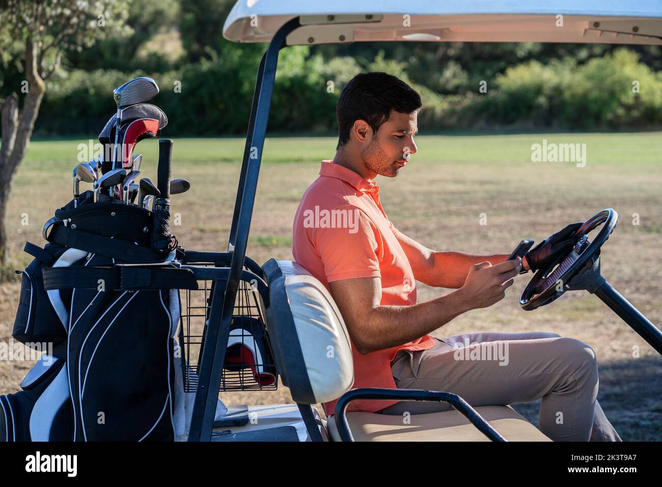 Messaggi di testo per gli atleti adulti di sesso maschile sul cellulare a bordo di un veicolo con attrezzatura da golf sul campo alla luce del sole Foto Stock