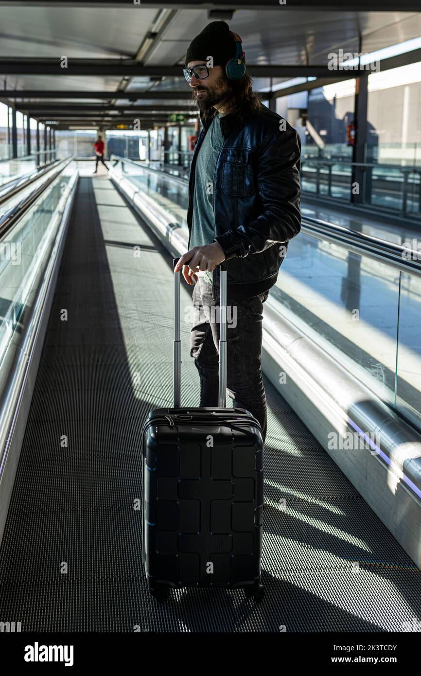 Uomo hipster in giacca di pelle nera e cappello lavorato a maglia camminando con valigia sul tapis roulant nel terminal dell'aeroporto Foto Stock