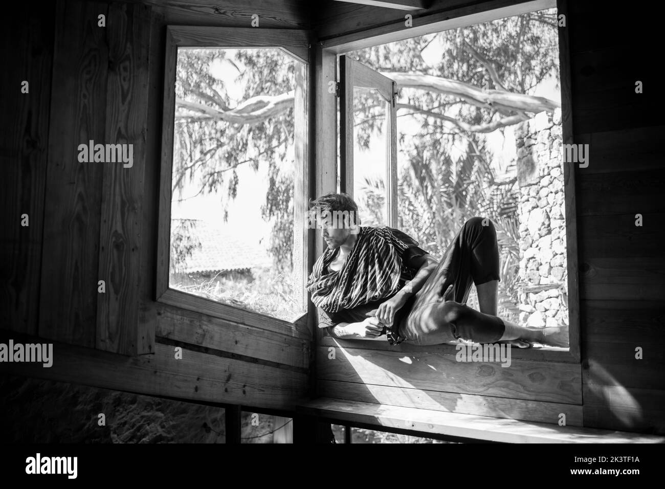 Giovane modello etnico maschile contemplativo in stile indossare guardando lontano dalla finestra di casa di campagna in legno Foto Stock