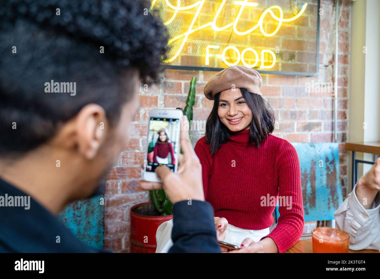 Uomo con smartphone che fotografa una felice ragazza etnica sorridente in un ristorante moderno Foto Stock