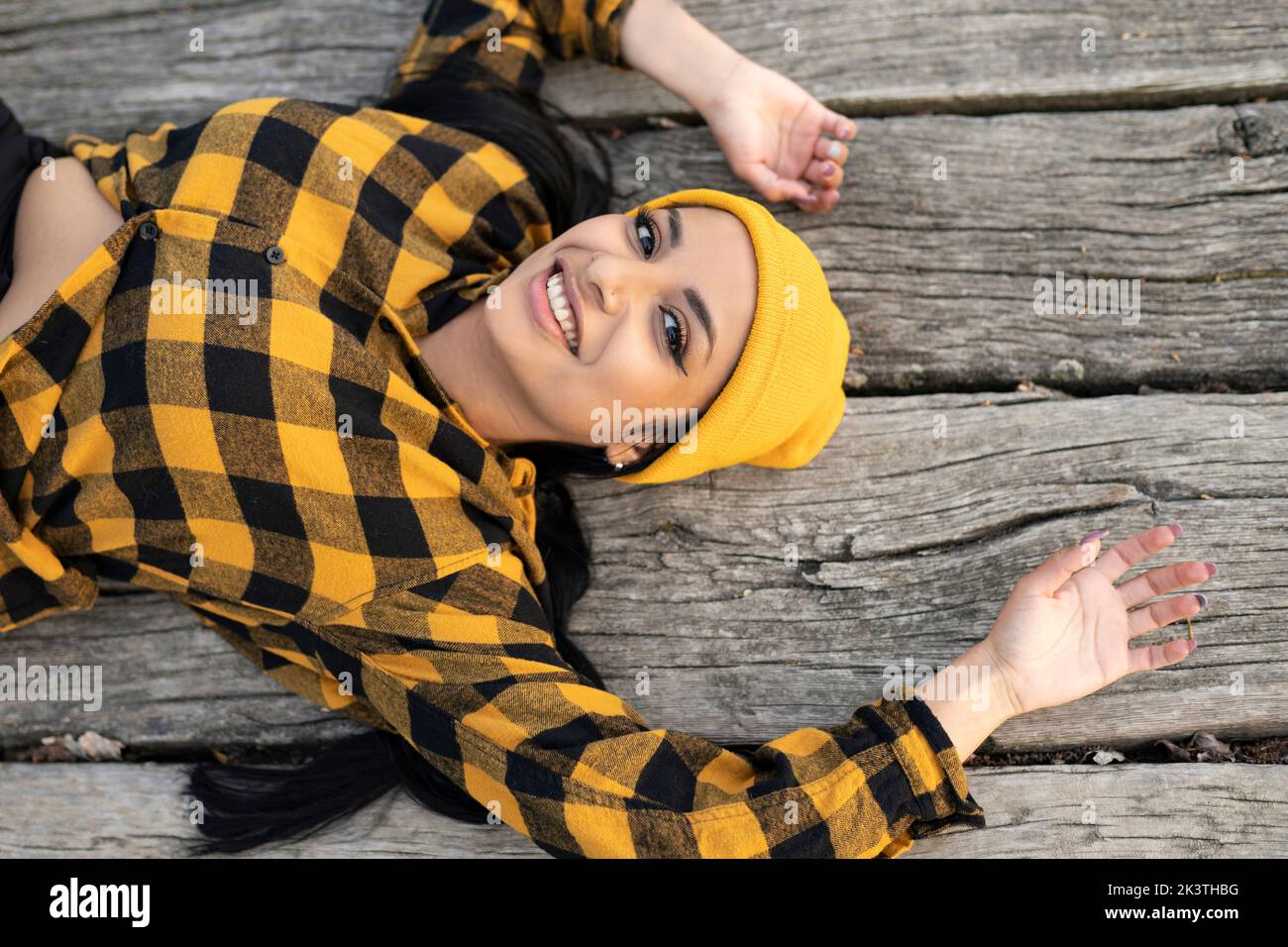 Vista dall'alto di una giovane ragazza latinoamericana sicura in una camicia a scacchi alla moda e un cappello giallo sorridente e guardando la macchina fotografica mentre sdraiato sul bordo di legno Foto Stock