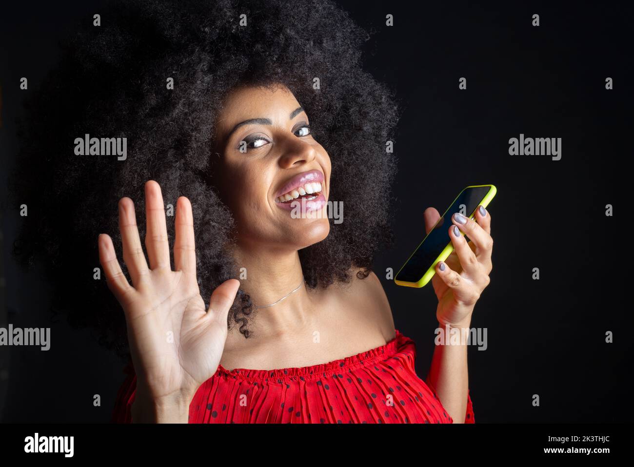 Giovane ragazza etnica allegra con cellulare e acconciatura afro dicendo ciao con mano sollevata mentre si guarda la macchina fotografica Foto Stock